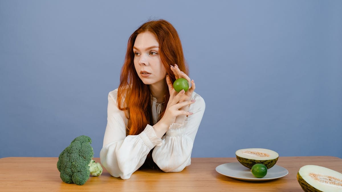 Caucasian woman sitting at table holding lime with broccoli and melon.
